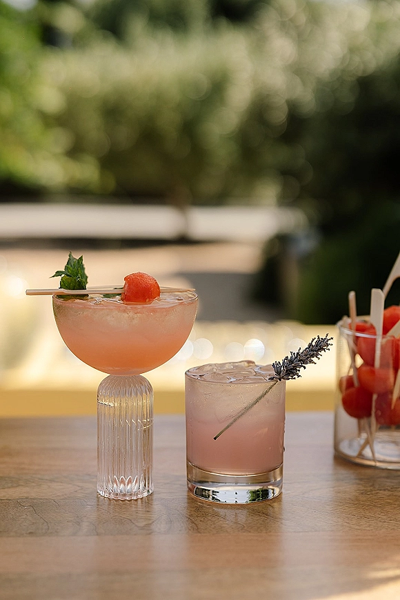 Wedding cocktails displayed as signature wedding drinks in coupe and rocks glasses with watermelon, mint, and lavender garnish on a wooden table outdoors