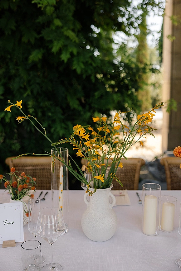 Reception tablescape with yellow floral centerpiece in a ceramic vase, pillar candles and glassware on white linen, set on a patio with trees