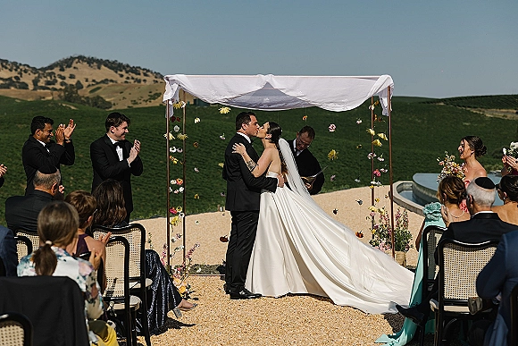 Wedding kiss as bride in satin dress and long veil embraces groom in tux under draped floral arch on terrace with hills beyond