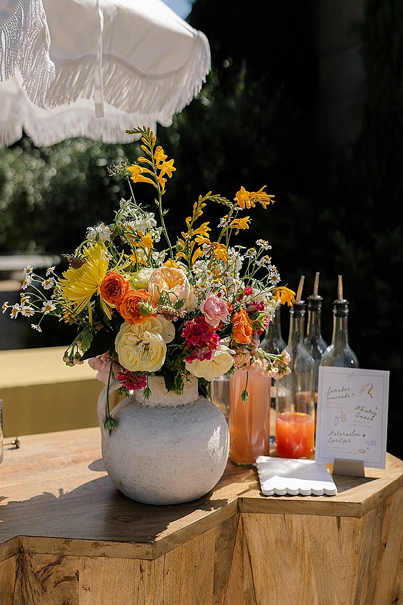 Wedding centerpiece with colorful wedding flowers in a ceramic vase on a wooden table, accented by an orange drink and bottles in sunlight