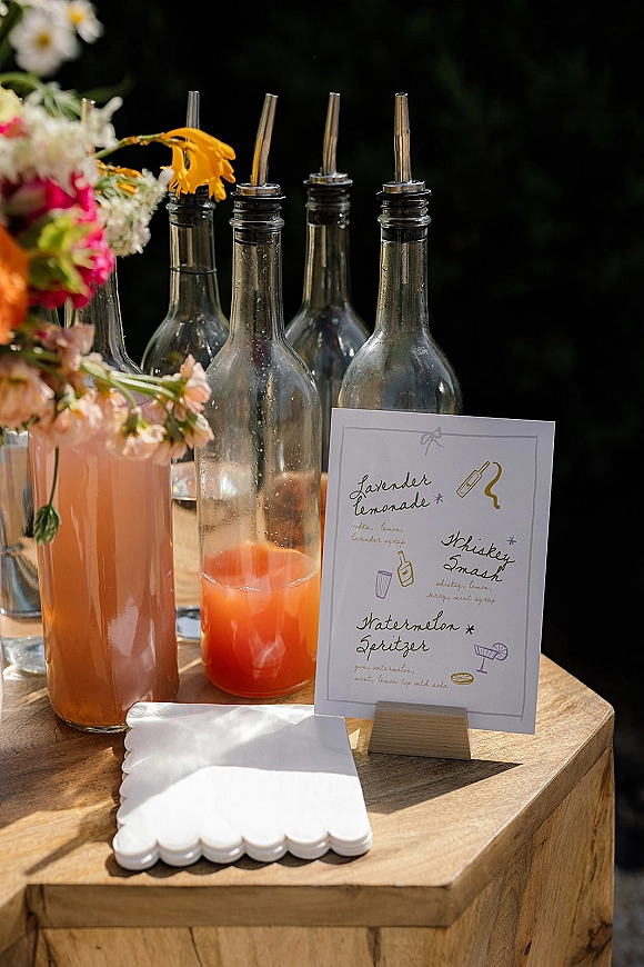 Wedding bar menu with signature drink menu on a wooden stand beside pour-spout bottles, mixers, and a small floral vase against dark greenery backdrop