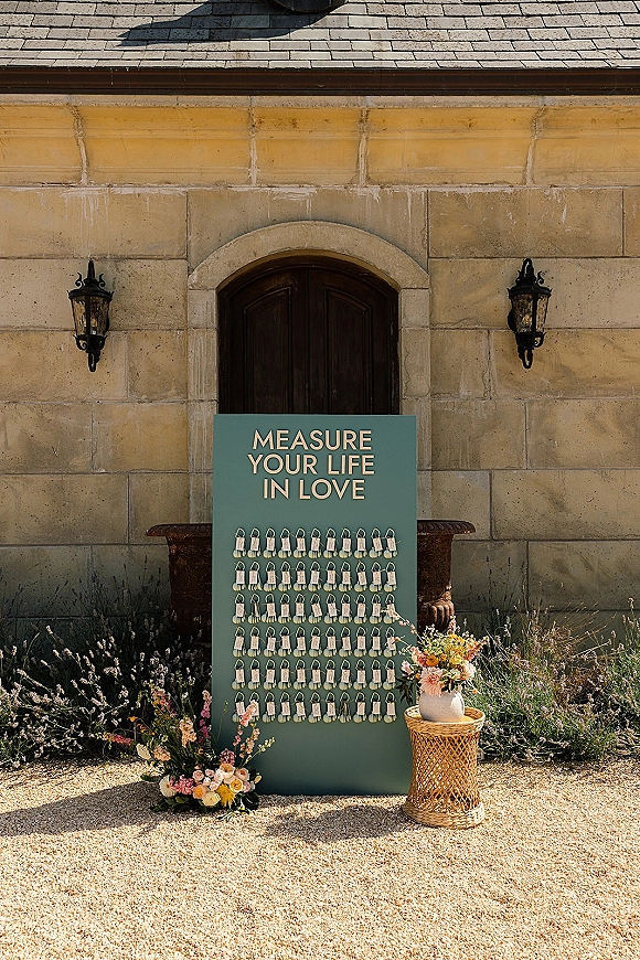 Wedding seating chart with escort card display on a green board with gold lettering, framed by florals on a wicker table by a stone wall door