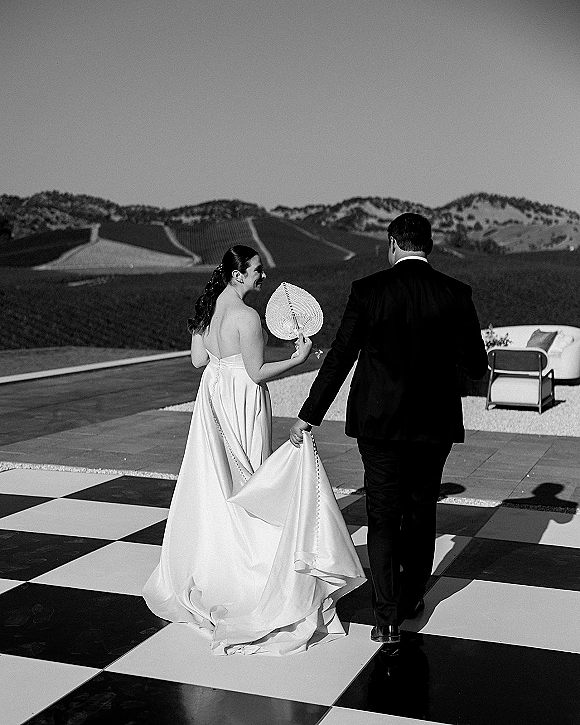 Couple portrait of newlyweds holding hands from behind, bride in veil and long train with hand fan on a patio with mountains beyond