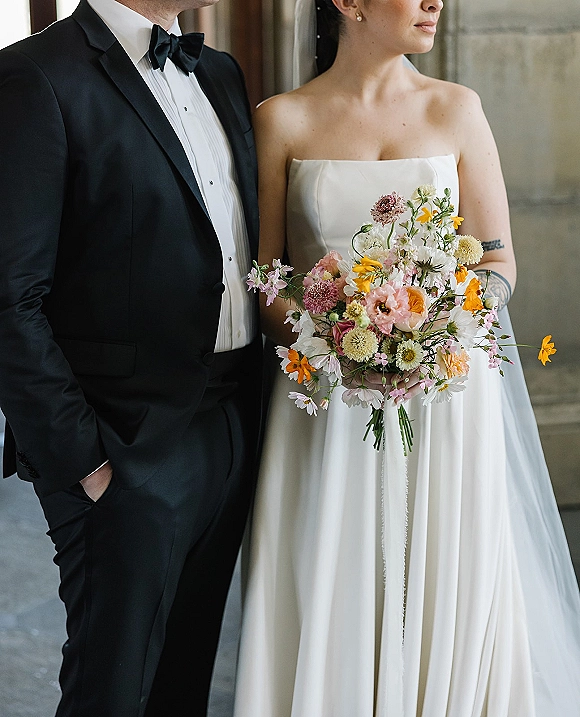 Couple portrait of bride in a strapless wedding dress holding a wildflower wedding bouquet beside groom in black tuxedo by stone wall doorway