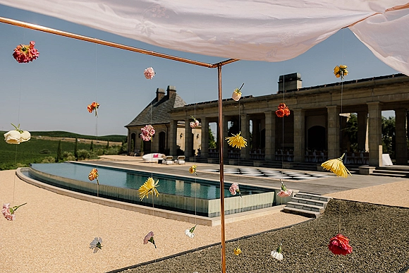 Outdoor wedding canopy with draped fabric on a copper frame, floral strings hanging above a poolside patio with stone villa backdrop