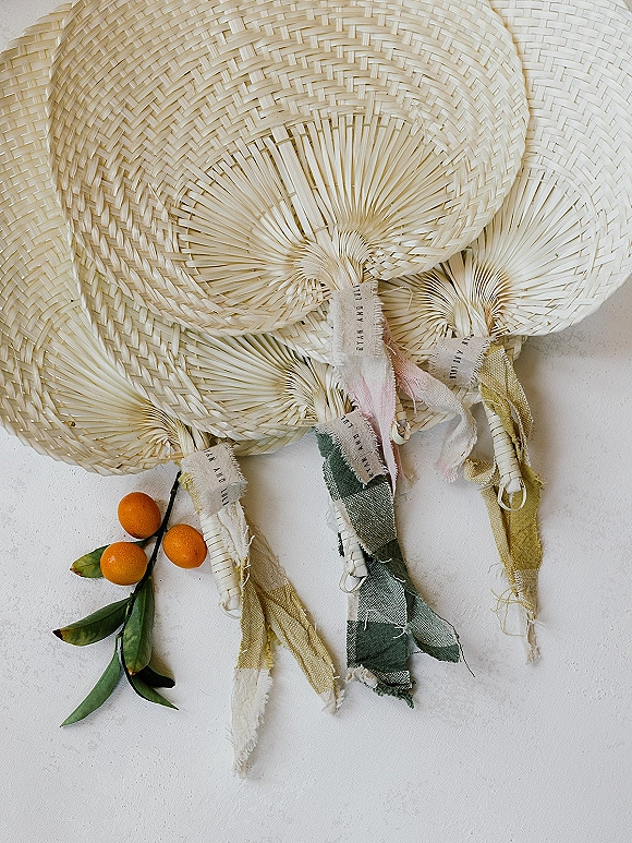 Wedding hand fans with woven palm leaf blades and fabric ribbons, accented by small oranges and leaves on a white textured surface