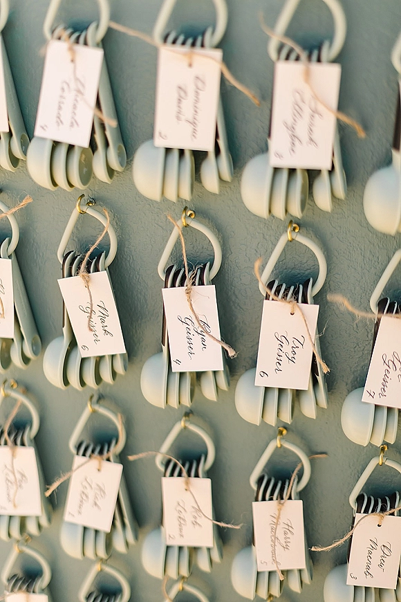 Wedding escort cards hanging as an escort card display, calligraphy name tags tied with twine to measuring spoons and rings on a painted wall