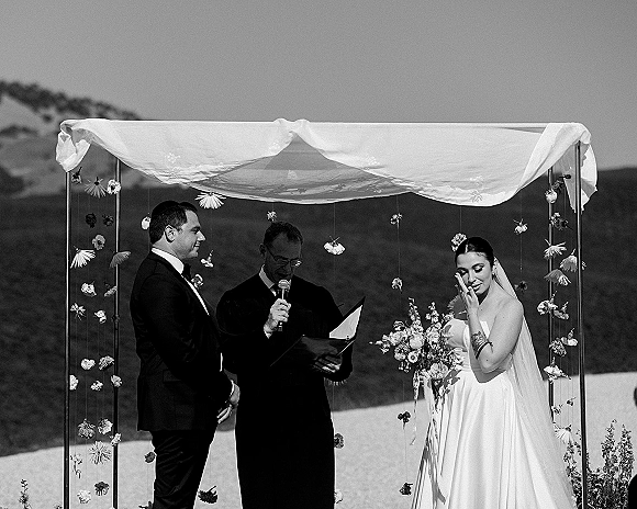 Wedding vows at an outdoor wedding ceremony as bride wipes tears beside groom in tuxedo under draped arch with mountains behind