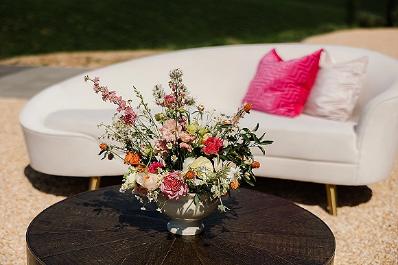 Wedding lounge seating with a white sofa and pink pillows around a round wooden coffee table topped with a floral centerpiece on gravel by lawn