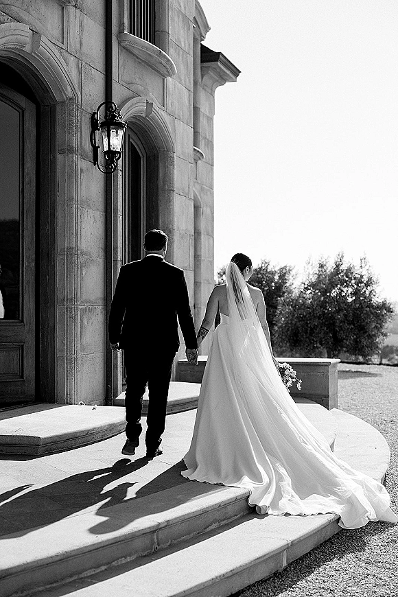 Couple portrait of bride and groom walking away holding hands, her long veil and bouquet trailing on stone steps by arched doorway