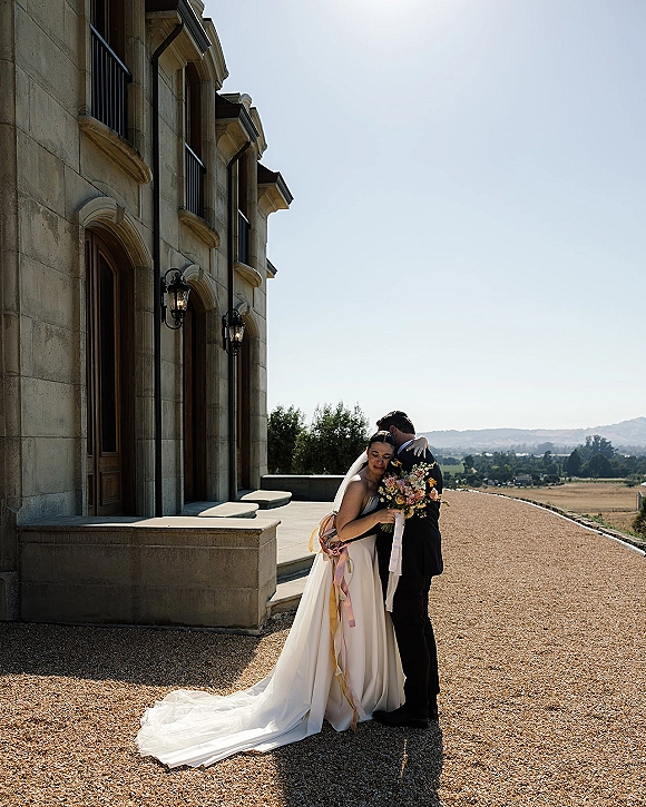Couple portrait of bride and groom embrace, her veil and wildflower bouquet with ribbon streamers beside a stone archway courtyard