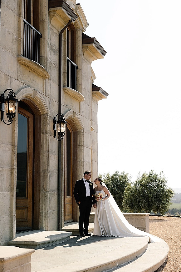 Couple portrait of bride and groom holding hands on outdoor stone steps by an arched doorway, her veil and bouquet catching light