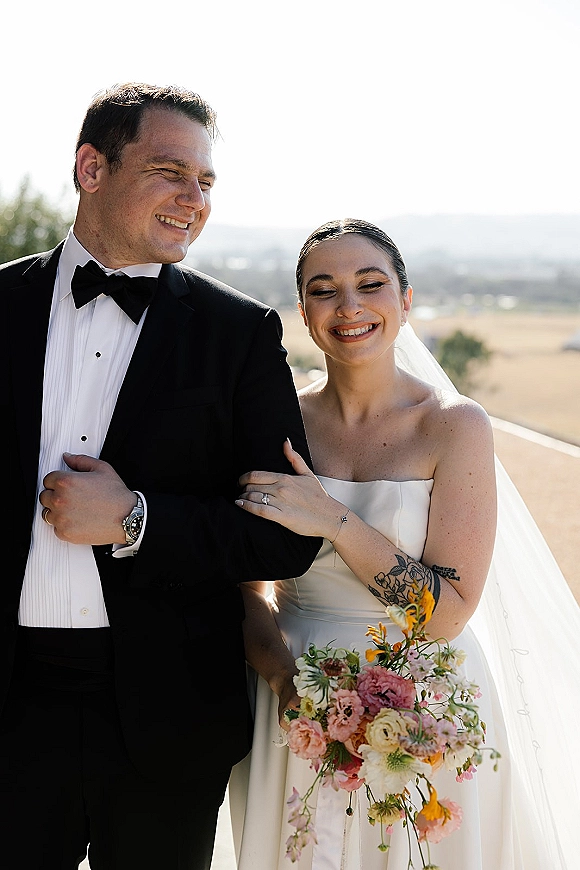 Couple portrait of bride holding bouquet beside groom in black tuxedo, her veil flowing against bright sky over rolling hills