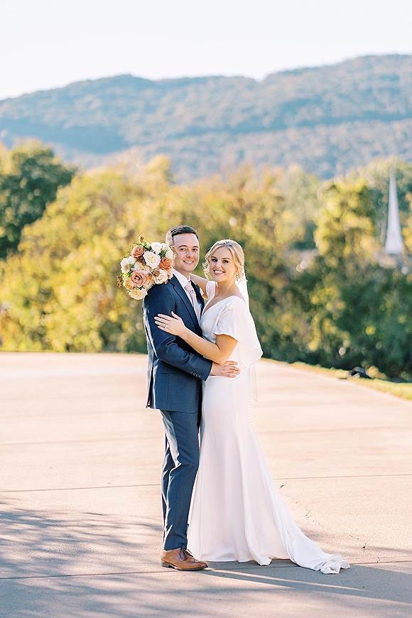 Couple portrait of bride and groom embracing, her rose bouquet and veil against mountains and a church steeple in soft light