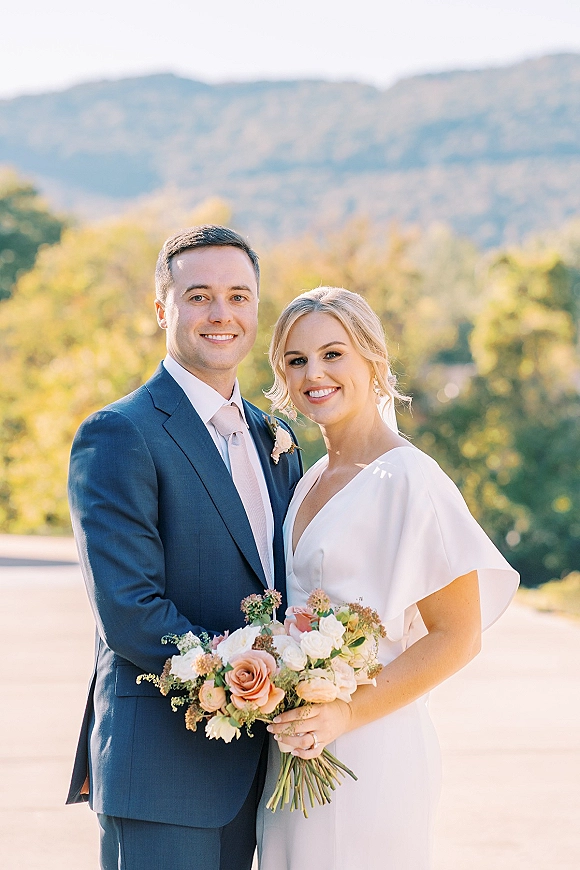 Couple portrait of bride and groom smiling, bride holding a white rose bouquet, with mountains and trees in the background