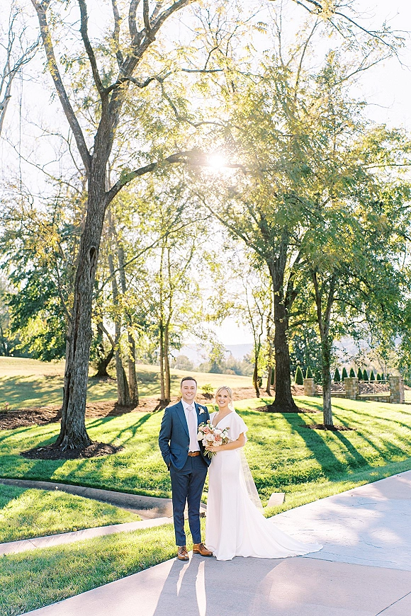 Couple portrait of bride holding a blush bouquet beside groom in a suit, standing on a sunlit lawn path with trees and hills behind