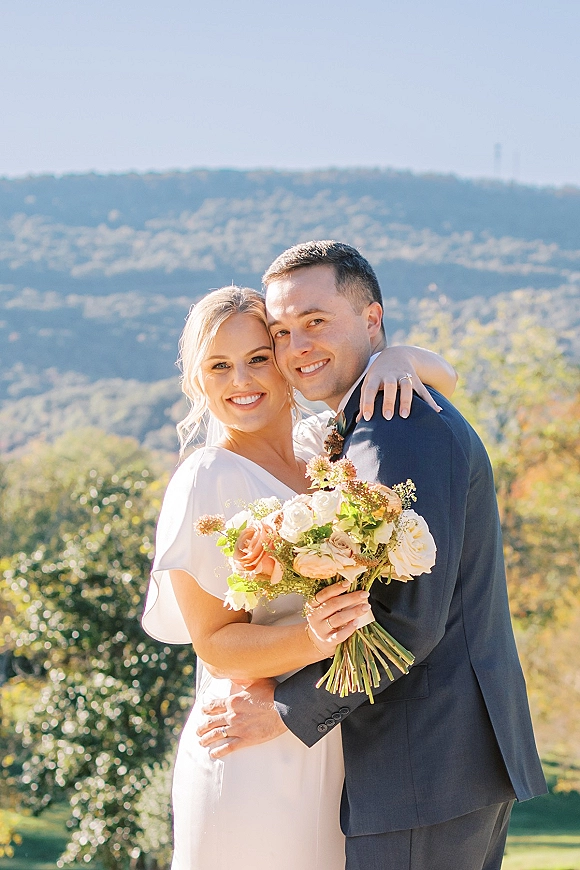 Couple portrait of bride and groom embrace, her veil and peach bouquet visible, posing on a sunny hillside with mountains and trees