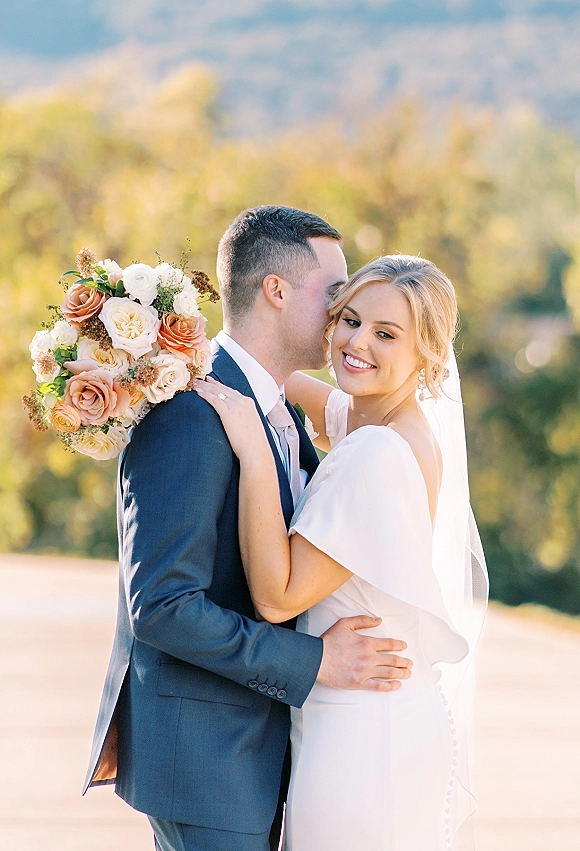 Couple portrait of groom kissing bride’s cheek as she smiles holding a rose bouquet, veil flowing, with hills and greenery behind