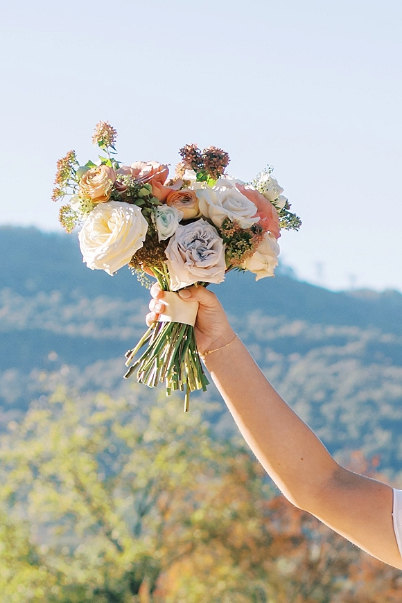 Bridal bouquet of pastel roses and greenery with ribbon wrap, held up against a mountain landscape with trees and blue sky