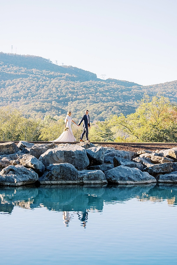 Couple portrait of bride and groom holding hands by a mountain lake, her veil trailing and bouquet in hand on a rocky shore