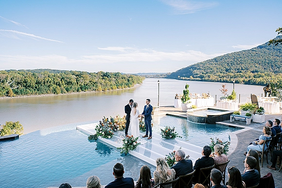 Wedding ceremony with an outdoor wedding ceremony arch as bride and groom stand with officiant on a terrace patio by pool, river and mountains
