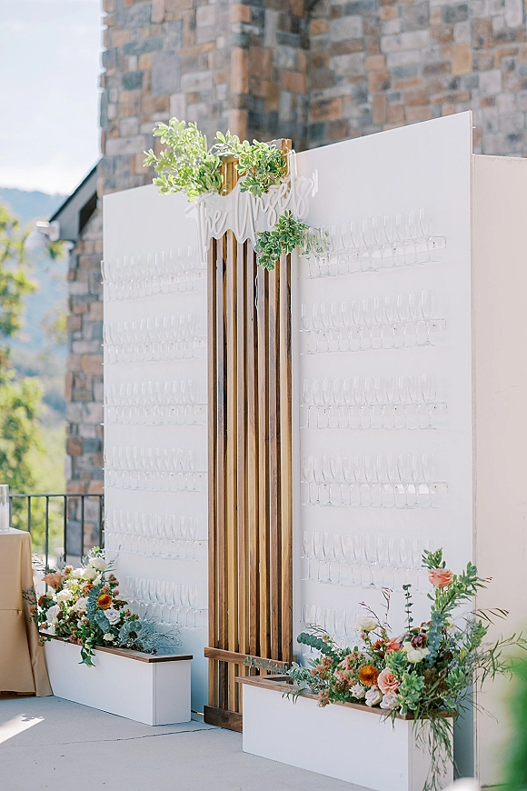 Champagne wall with wedding champagne wall flutes and an acrylic sign, framed by greenery and florals on a mountain terrace backdrop