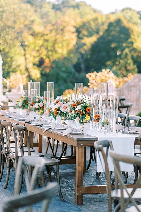 Reception tablescape with an outdoor wedding reception table on a wood farm table, floral centerpieces, candlelight, and crossback chairs on a patio under trees