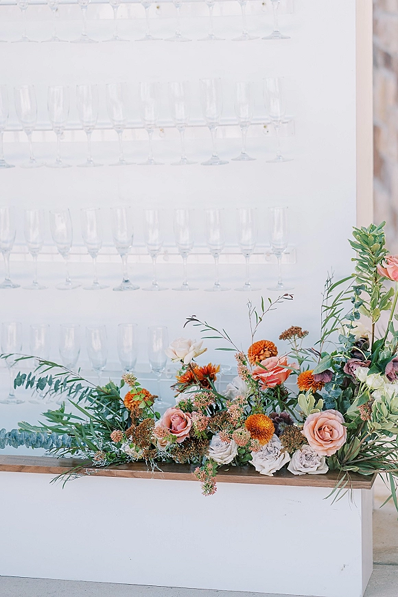 Champagne wall with rows of champagne flutes and a rose-and-eucalyptus accent on a modern white display wall for cocktail hour
