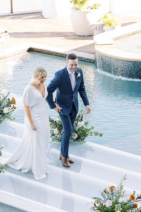 Wedding couple portrait of bride and groom walking hand in hand poolside, her long veil flowing as they descend steps by water feature