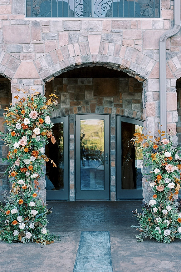 Wedding ceremony arch with asymmetrical floral arch columns of roses and greenery framing a stone entryway with glass double doors