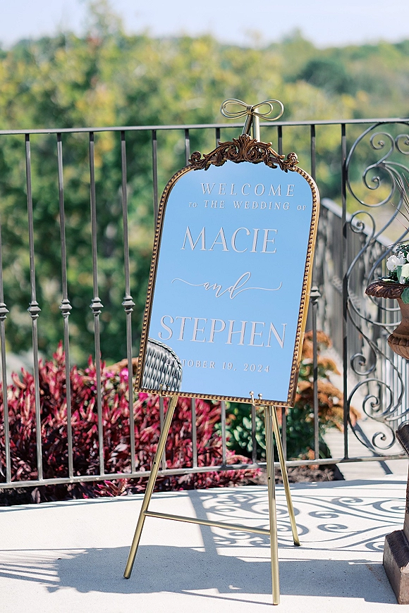 Wedding welcome sign in a gold framed ornate frame on a gold easel with ribbon bow and floral urn on an outdoor terrace by railing