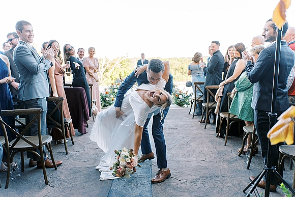 Recessional moment as newlyweds walk the wedding recessional aisle, bride in gown and veil holding bouquet, guests cheering on terrace with hills beyond