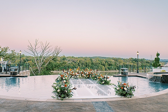 Ceremony setup with a clear aisle wedding leading to a floral arch, poolside on a stone patio with fountains and river view