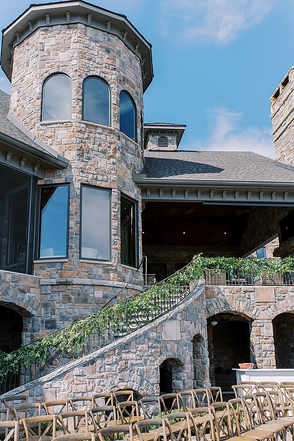 Wedding venue exterior with greenery garland on a wrought iron railing, stone building with arched windows, tower and patio staircase under blue sky