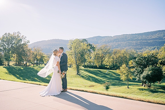 Wedding couple portrait of the bride and groom kissing, her veil trailing and bouquet in hand, on a mountain path under blue sky