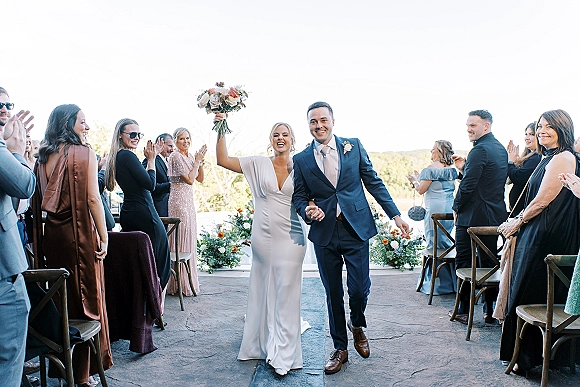 Wedding recessional with bride and groom walking aisle, holding hands as she lifts bouquet overhead, guests cheering on terrace with hills behind