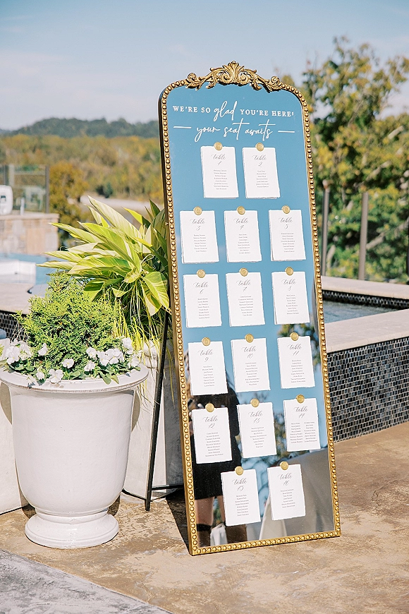 Wedding seating chart mirror in a gold ornate frame with blue backing and pinned cards on a black easel by a poolside patio