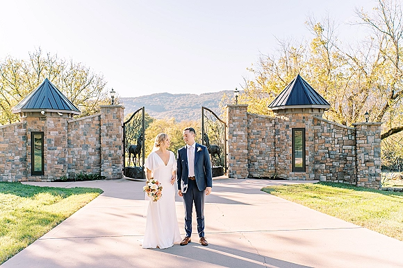 Couple portrait of bride and groom portrait holding hands by a stone gate, bride with a bouquet beside wrought iron gates and hills