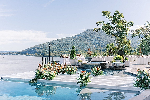 Reception setup with round tables in white linens and floral centerpieces on a terrace by the lake, mountains beyond and a pool nearby