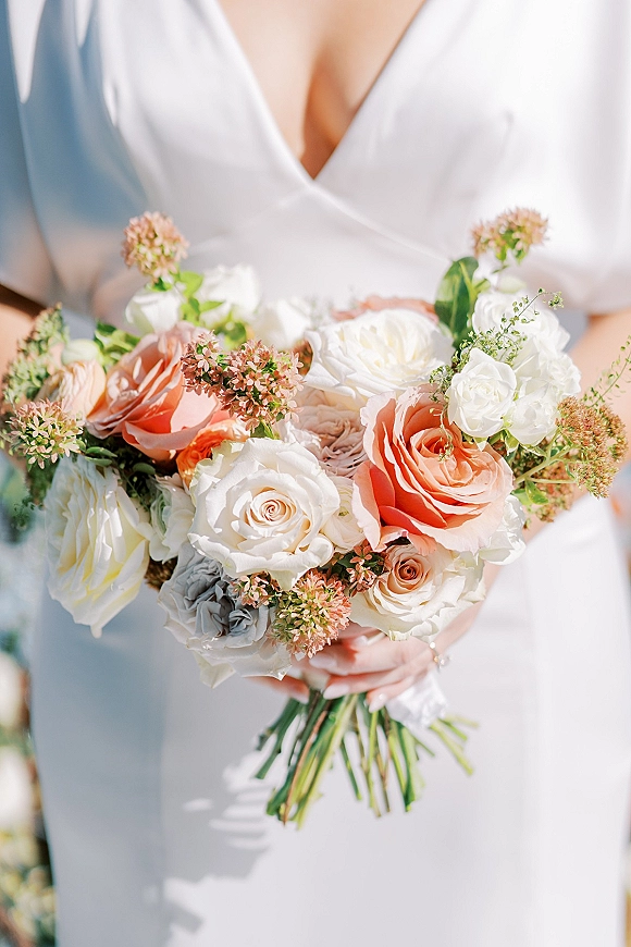 Bridal bouquet of peach and white bouquet roses with pink filler flowers and greenery, ribbon-wrapped stems against a soft blurred backdrop