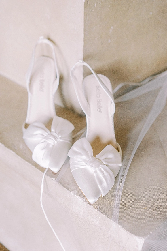 Bridal shoes, white satin wedding heels with oversized bows and tulle veil draped over them on stone steps by a neutral wall