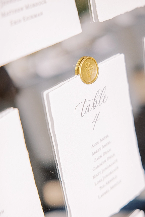 Wedding table number card in a clear acrylic holder with a gold wax seal beside a place card, set indoors with blurred signage