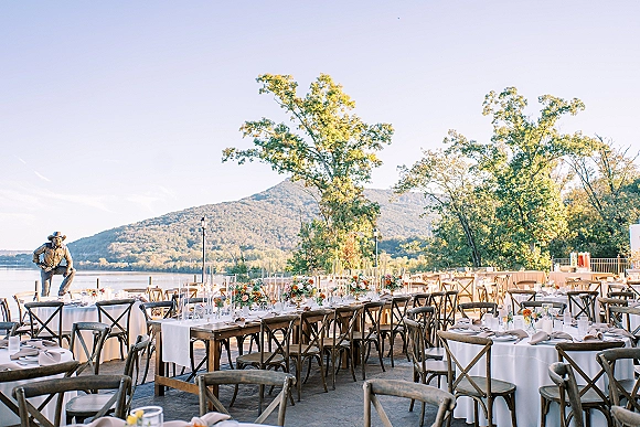 Outdoor reception setup with white linen round and banquet tables, floral centerpieces and candles on a lakeside terrace with mountains