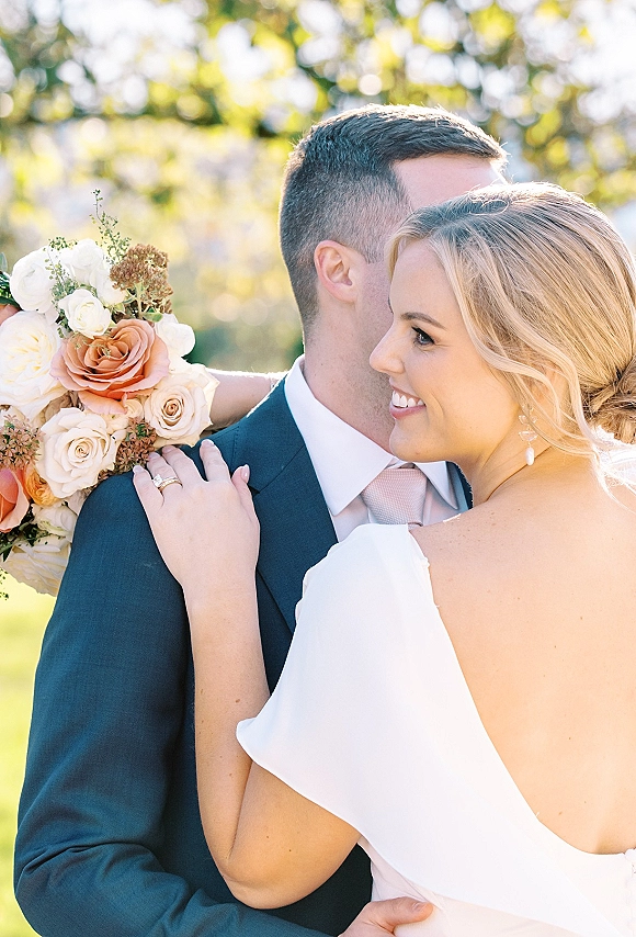 Couple portrait of bride and groom hug, bride smiling with peach rose bouquet and greenery against sunlit trees bokeh background