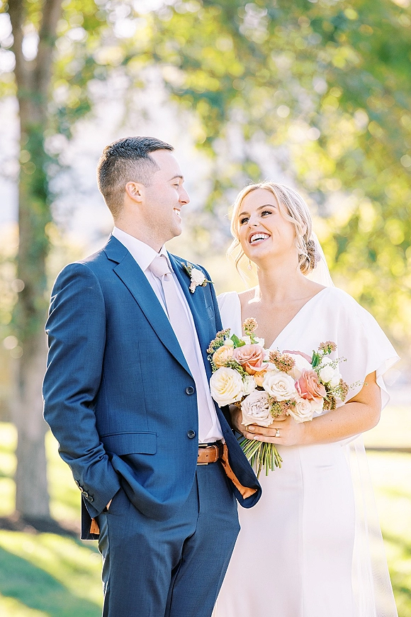 Couple portrait of bride and groom smiling, bride holding a bouquet in golden sunlight on a green lawn with trees behind