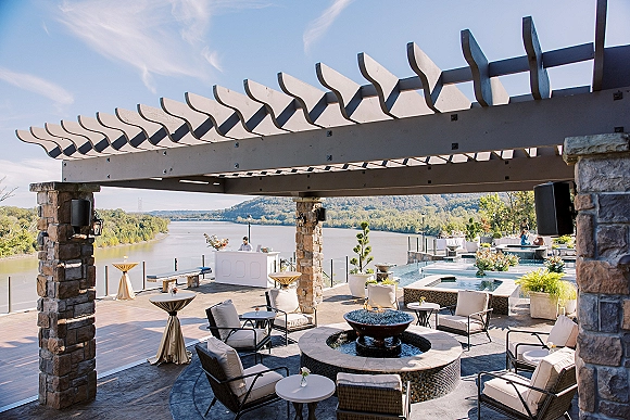 Outdoor reception lounge with wedding lounge seating under a stone pergola, sofas and fire pit by the pool overlooking a river and hills