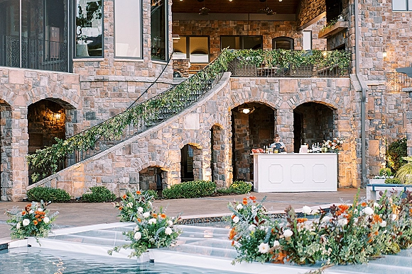 Outdoor wedding bar with a white portable wedding bar setup, bottles and glassware, plus floral accents by a poolside stone courtyard staircase