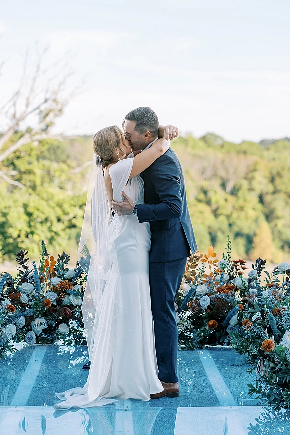 Wedding kiss portrait of bride and groom kissing, her long veil flowing as they embrace on a pool deck with trees and sky behind