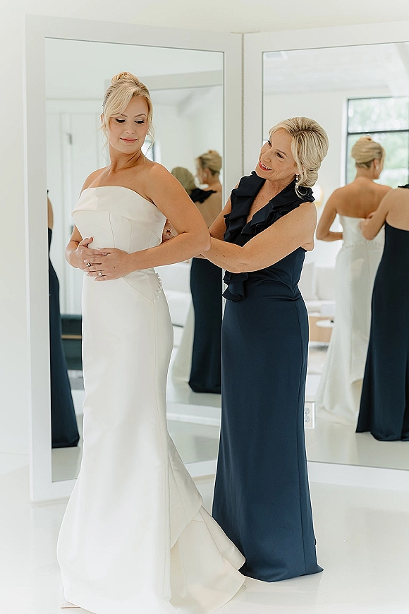 Wedding dress fitting as bride in a strapless button-back gown adjusts earrings and ring by mirrors in a bright bridal suite