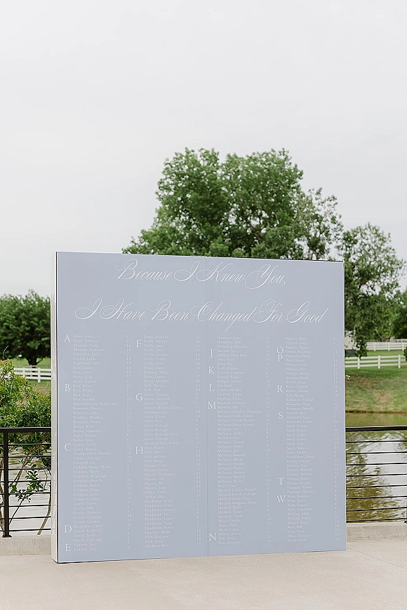 Seating chart sign with wedding seating chart calligraphy lettering on a large foam board, set by trees and a white fence near a pond